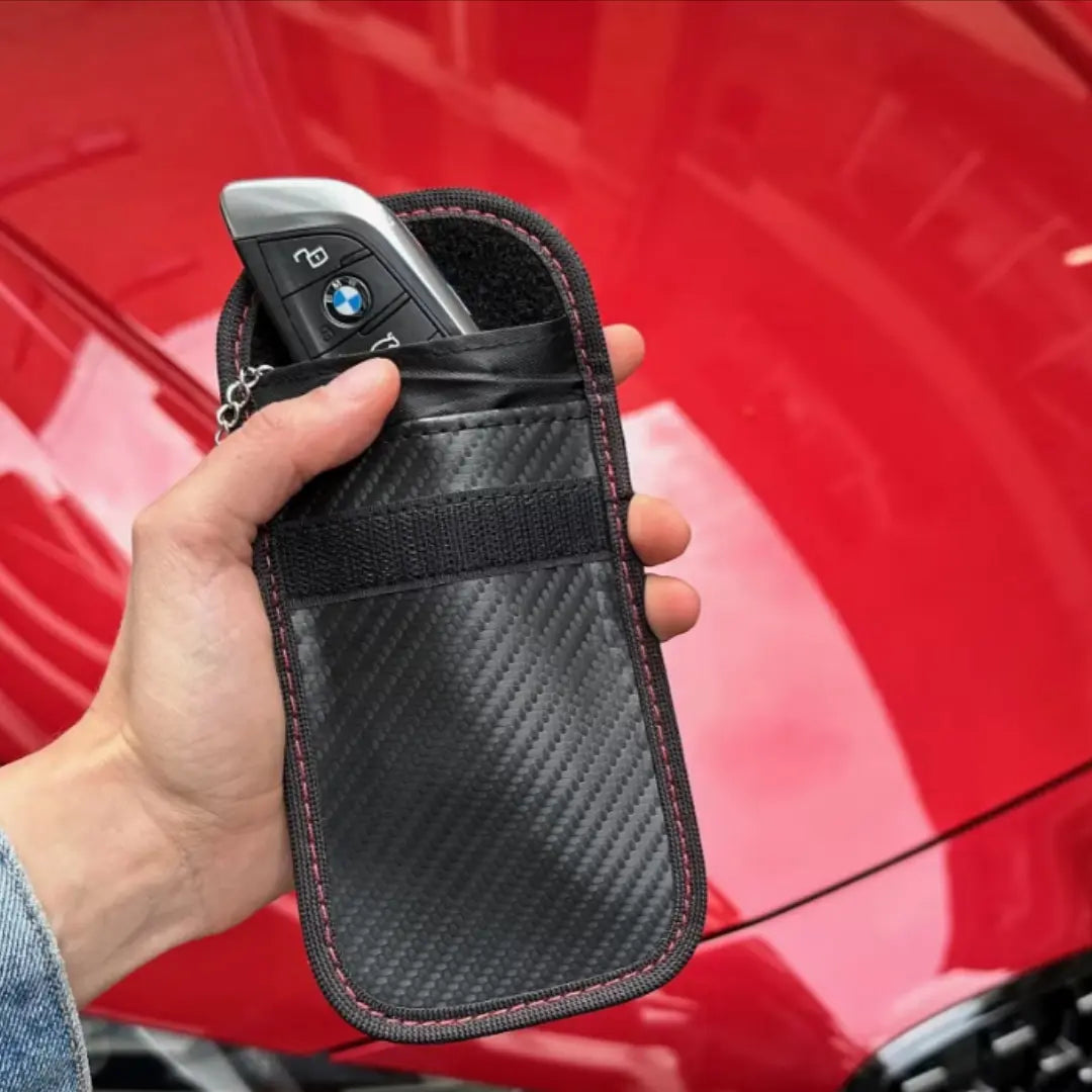 Hand holding a car key with a carbon fiber signal blocking faraday pouch in front of a red car.