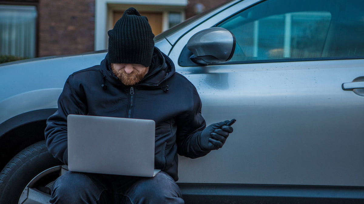 Person in dark clothing using a laptop and phone next to a car to conduct a signal relay attack